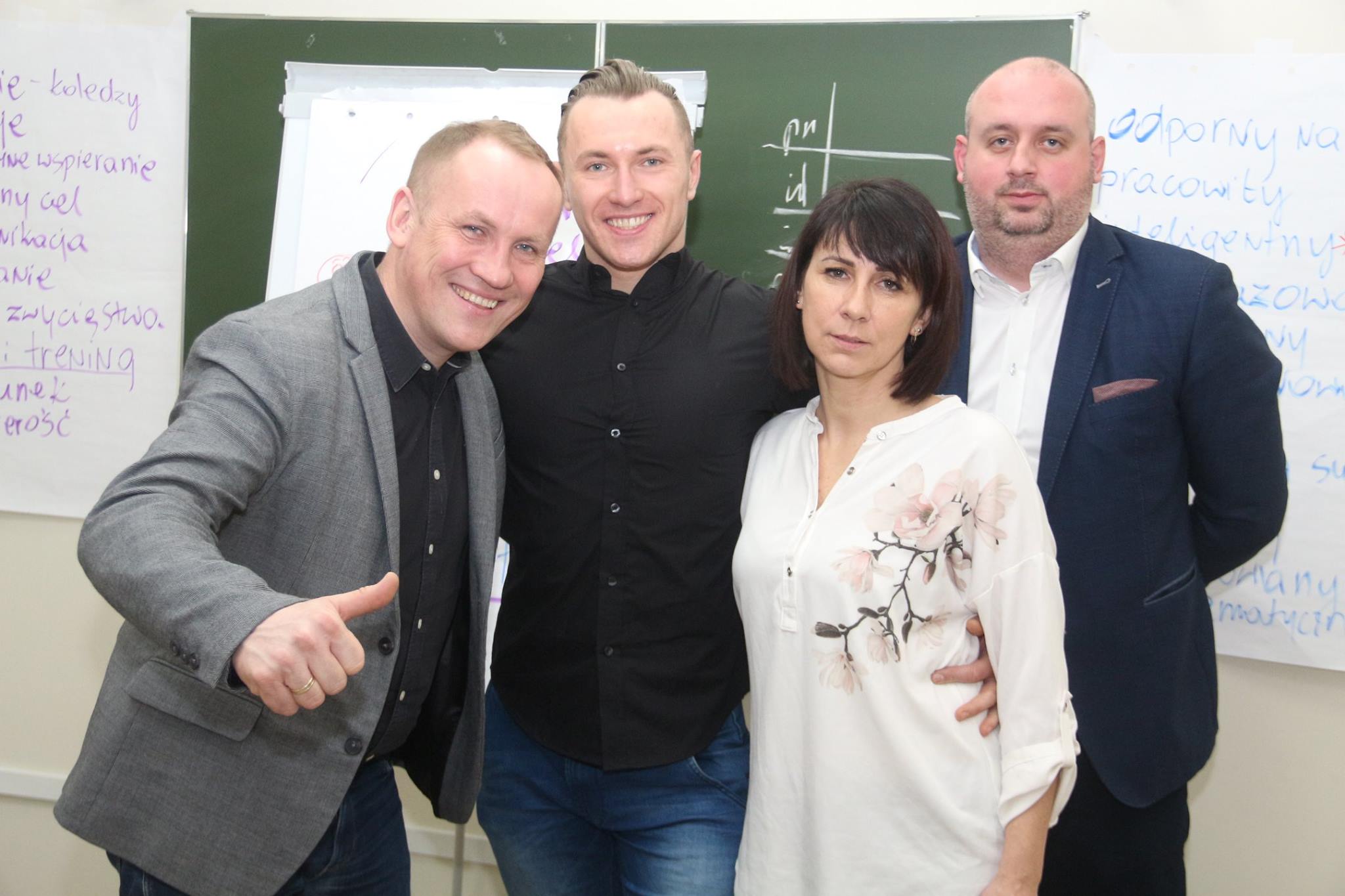 Cztery osoby w biurze pozują do zdjęcia, w tle szkoła. Trzy z nich uśmiechają się, a jedna ma poważny wyraz twarzy.