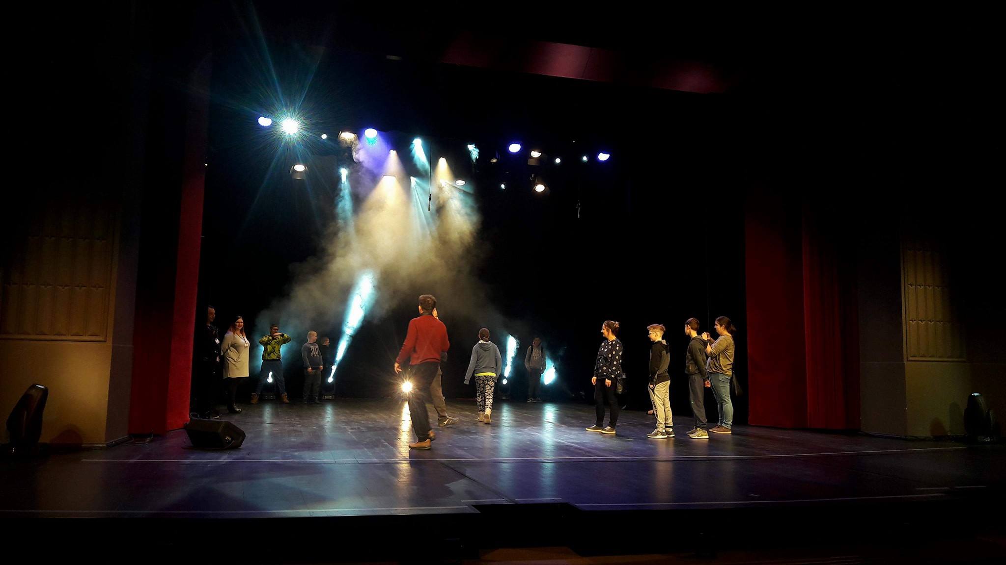 Grupa ludzi na scenie w oświetlonym teatrze, wśród efektów świetlnych i dymu.