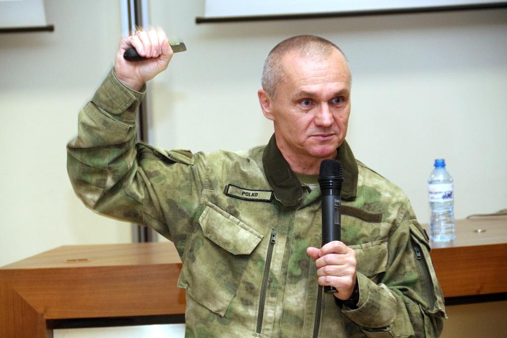 Mężczyzna w kamuflażowym mundurze wojskowym, trzymający mikrofon i wskazujący na coś, prowadzi prezentację.