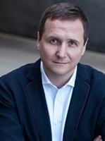 Mężczyzna w eleganckim garniturze, z poważnym wyrazem twarzy, siedzący w pomieszczeniu.