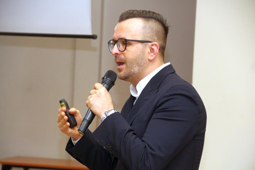 Mężczyzna w garniturze wygłasza przemówienie, trzymając mikrofon i pilot.