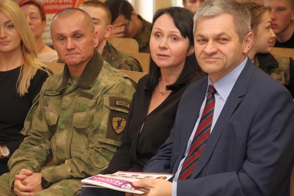 Grupa ludzi uczestniczy w wydarzeniu, siedząc na sali, z osobą w mundurze wojskowym na pierwszym planie.