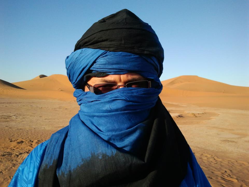 Mężczyzna w niebieskim turbanie stoi na pustyni, w tle wznoszące się wydmy.