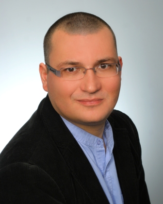 Mężczyzna w okularach, ubrany w ciemną marynarkę i jasną koszulę, z poważnym wyrazem twarzy.