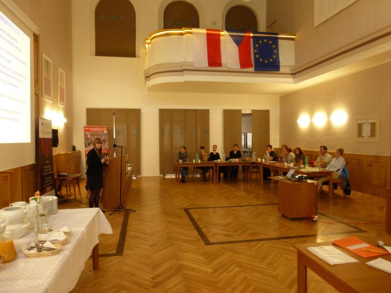 Wnętrze sali konferencyjnej z osobą mówiącą z podium, uczestnicy siedzą przy stołach. Na ścianie wiszą flagi.