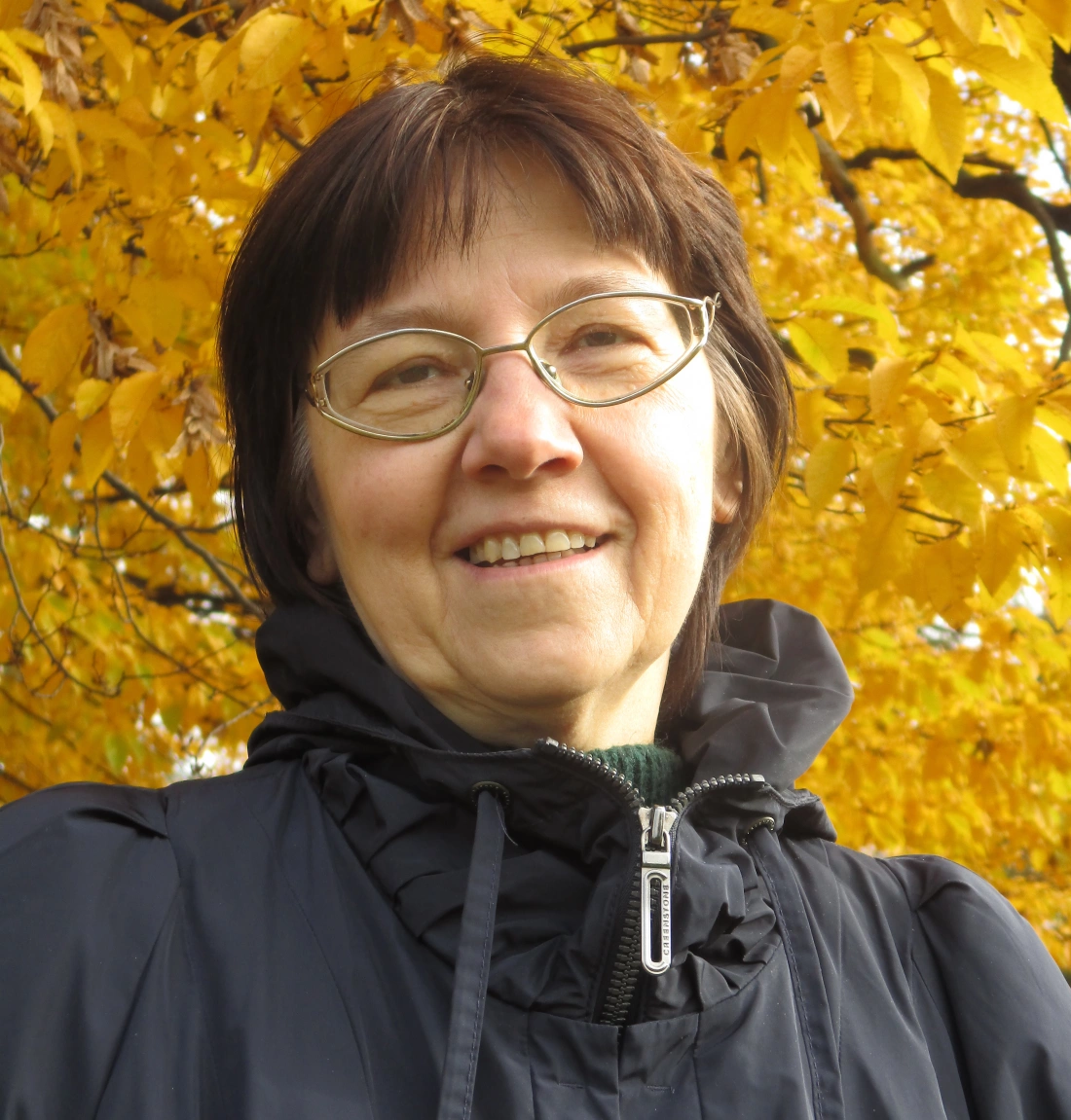 Uśmiechnięta kobieta w czarnej kurtce stoi przed żółtymi liśćmi drzew.