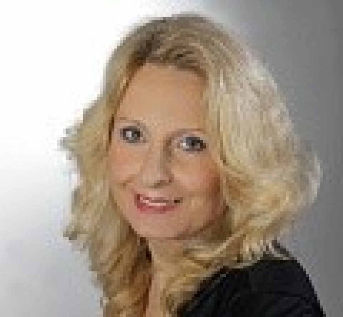 Kobieta z długimi, blond włosami, uśmiechająca się na tle szarej ściany.