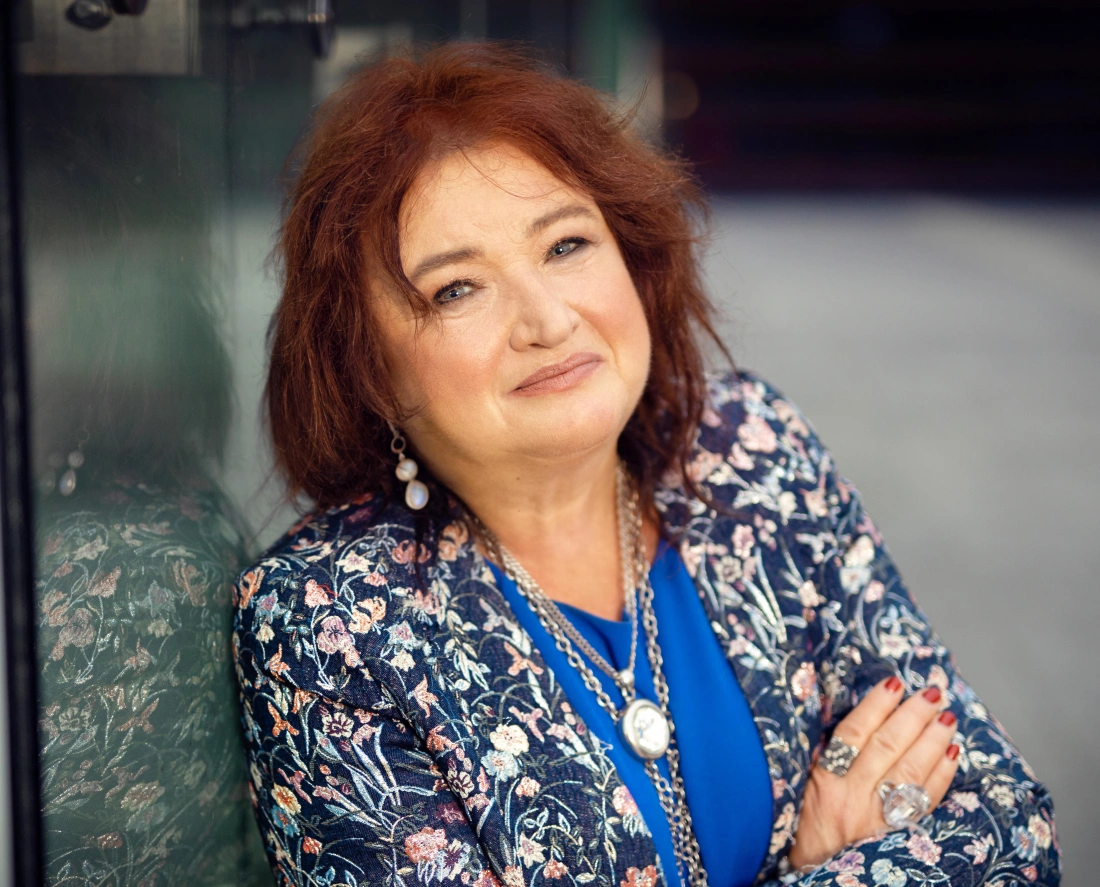 Kobieta o rudych włosach, uśmiechająca się, ubrana w kwiecisty żakiet i niebieską bluzkę, opiera się o szklaną powierzchnię.