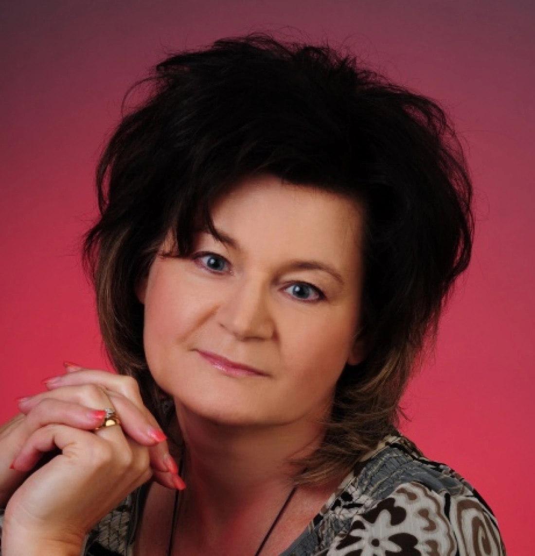 Portret kobiety z długimi, ciemnymi włosami, uśmiechającej się i spoglądającej w kamerę, na tle w odcieniach różu.