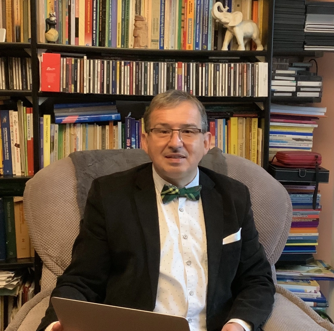 Mężczyzna w marynarce i muszce siedzi na fotelu z laptopem, w tle regał pełen książek.