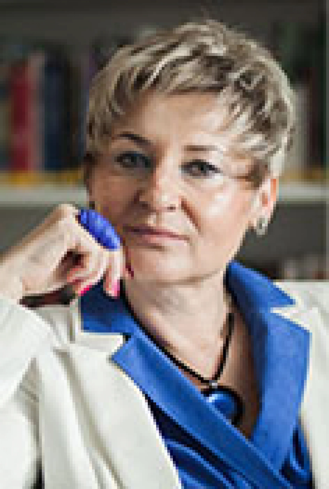 Kobieta z krótkimi, jasnymi włosami, uśmiechająca się, w niebieskim płaszczu, siedząca w stylowym wnętrzu.