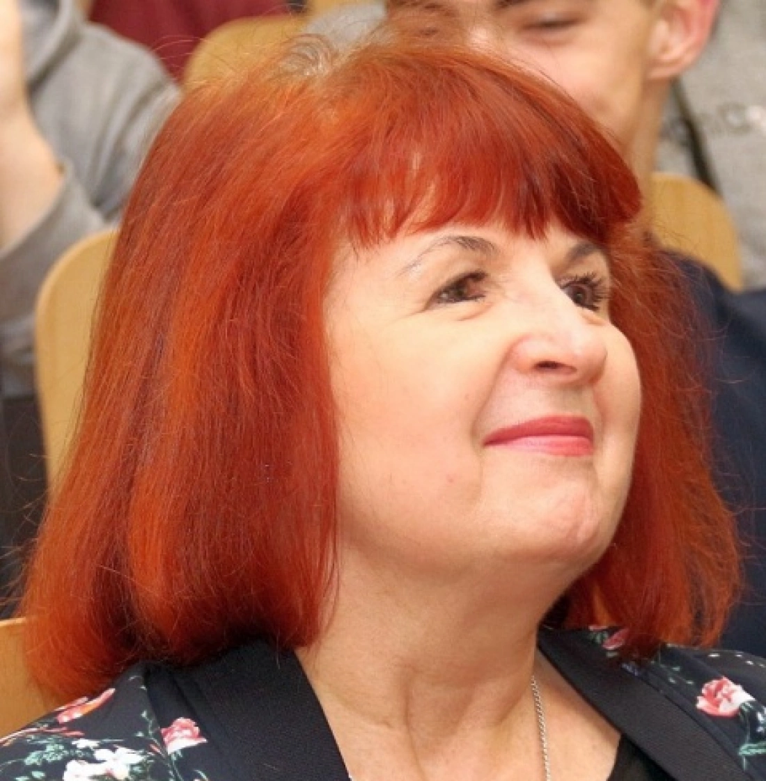 Kobieta z krótkimi rudymi włosami uśmiecha się, siedząc wśród innych osób.