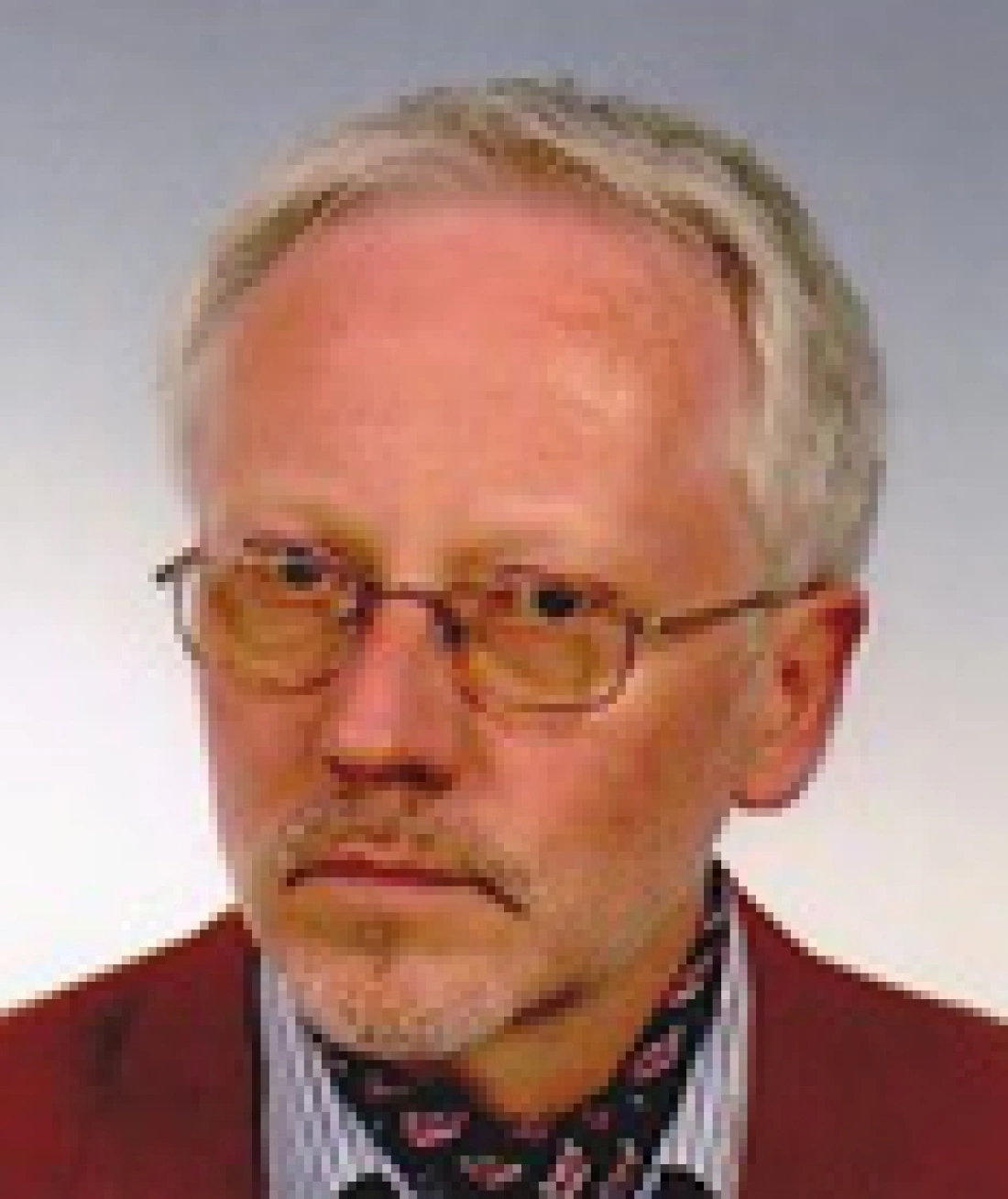 Mężczyzna z krótkimi, siwiejącymi włosami, w okularach i z poważnym wyrazem twarzy, ubrany w czerwony sweter i koszulę w paski.