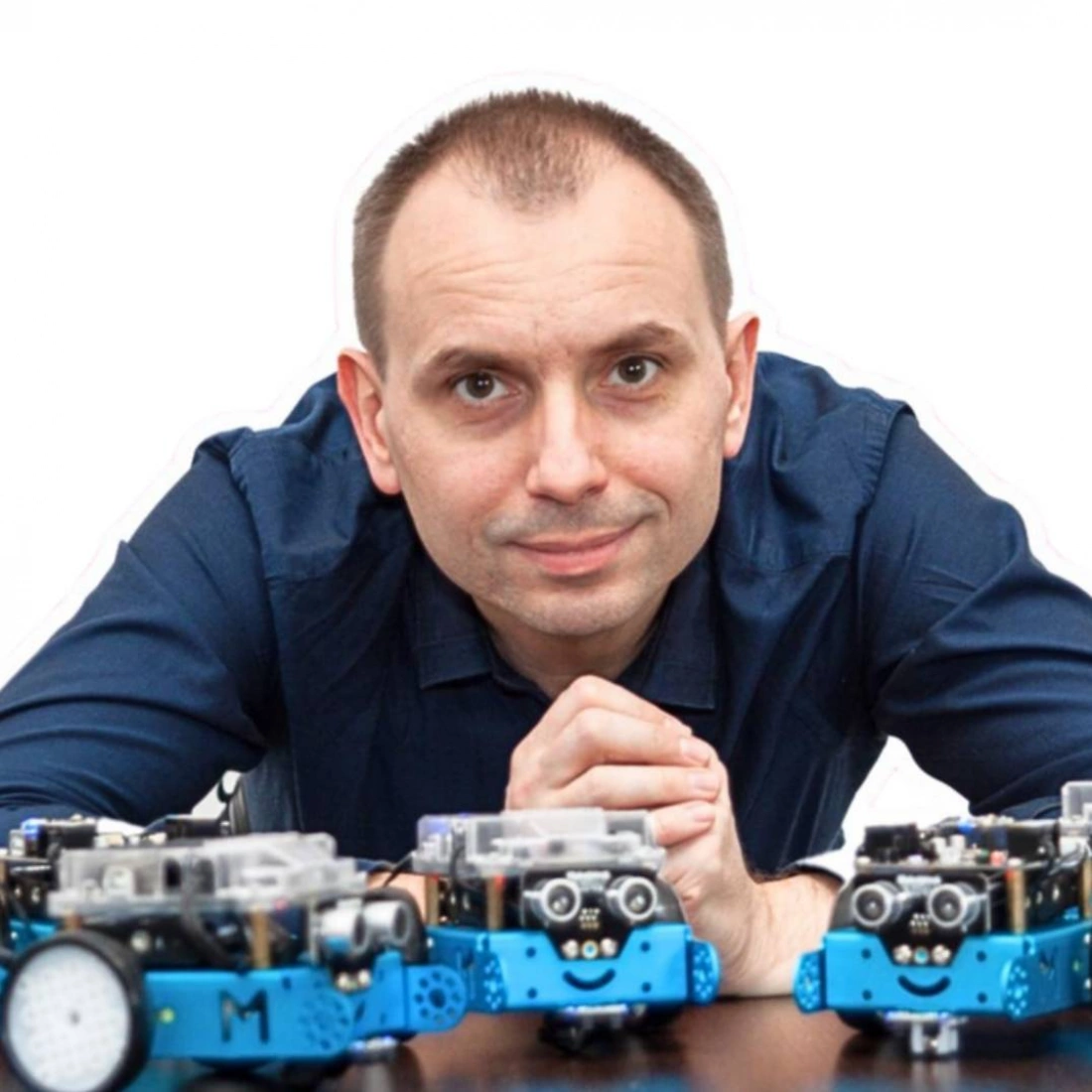 Mężczyzna w niebieskiej koszuli siedzi przed kilkoma robotami na stole.