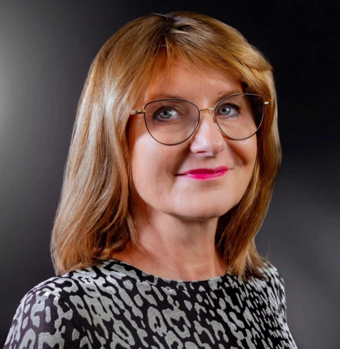 Portret kobiety z długimi, blond włosami, w okularach i uśmiechem, noszącej wzorzystą bluzkę.