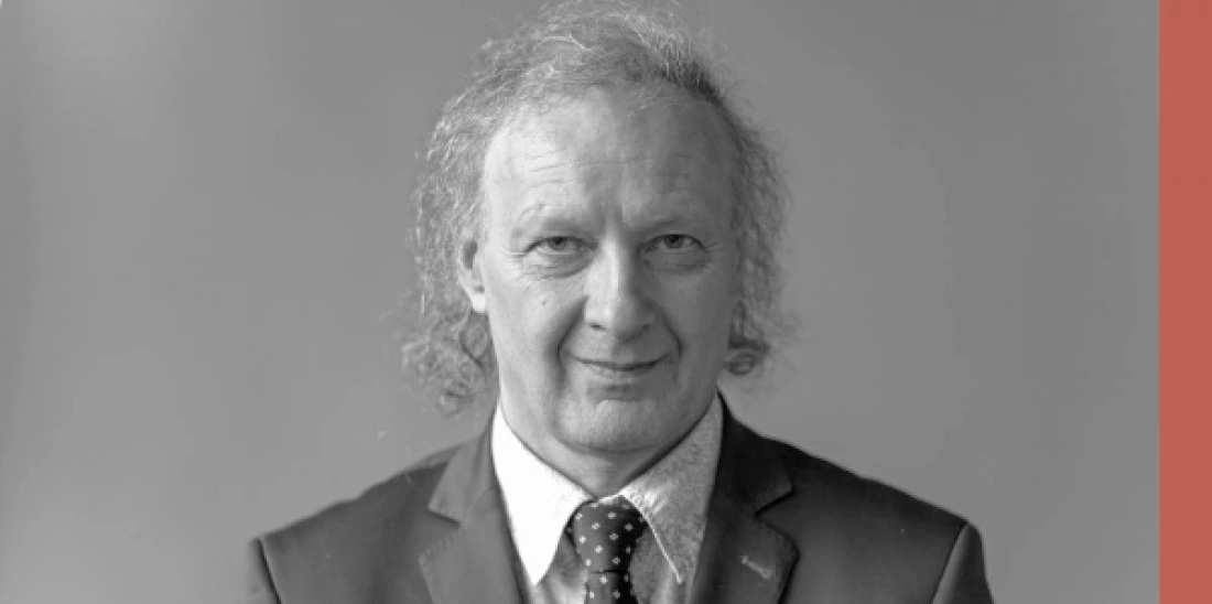 Mężczyzna w garniturze, z lekko kręconymi włosami, uśmiecha się na tle w jednolitym kolorze.