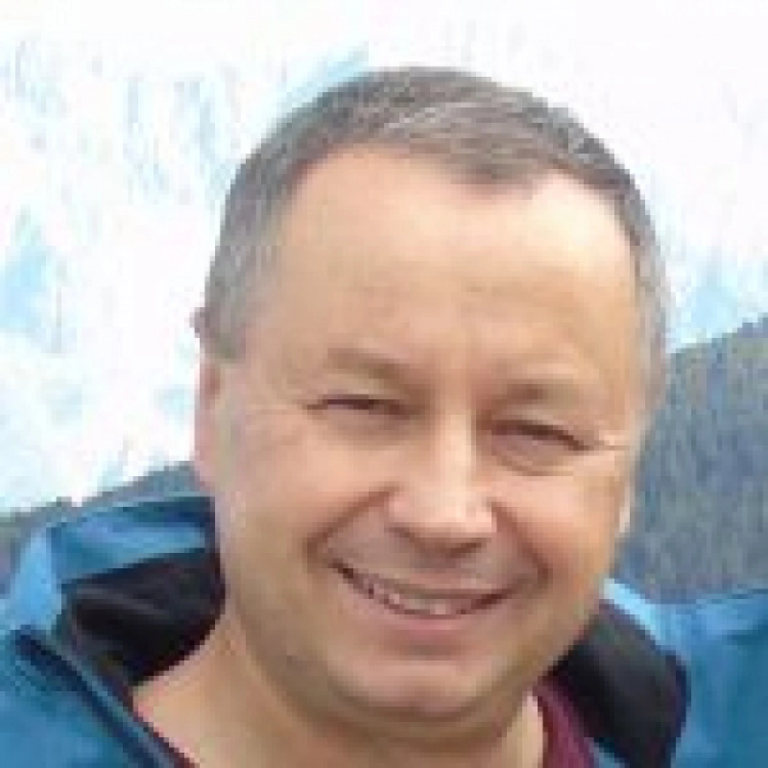 Mężczyzna uśmiechający się w niebieskiej kurtce z tłem górskim.