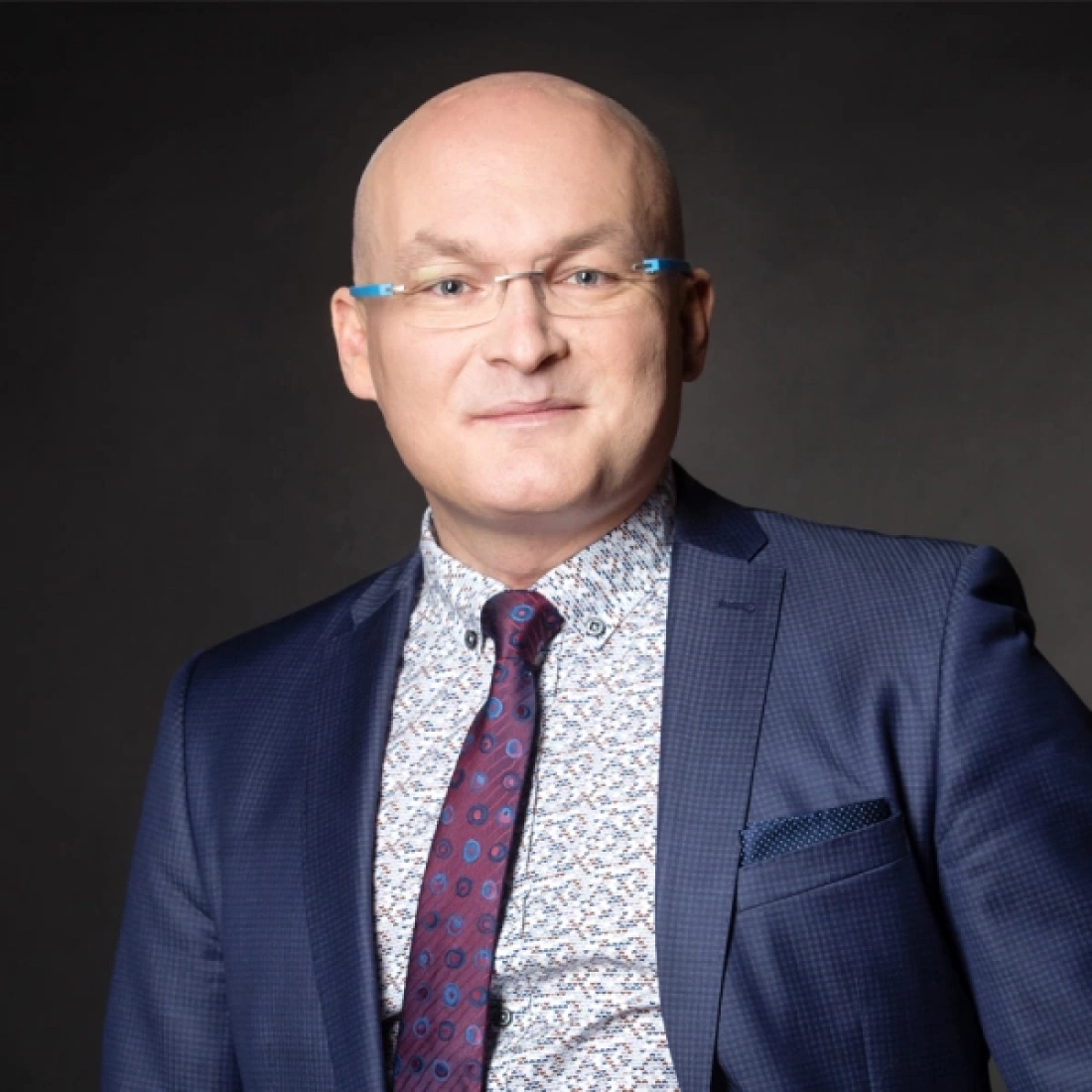 Mężczyzna w garniturze, z okularami, uśmiecha się do kamery.