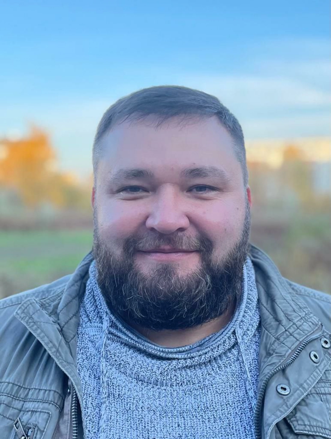 Mężczyzna z brodą uśmiechający się na tle nieba i natury.