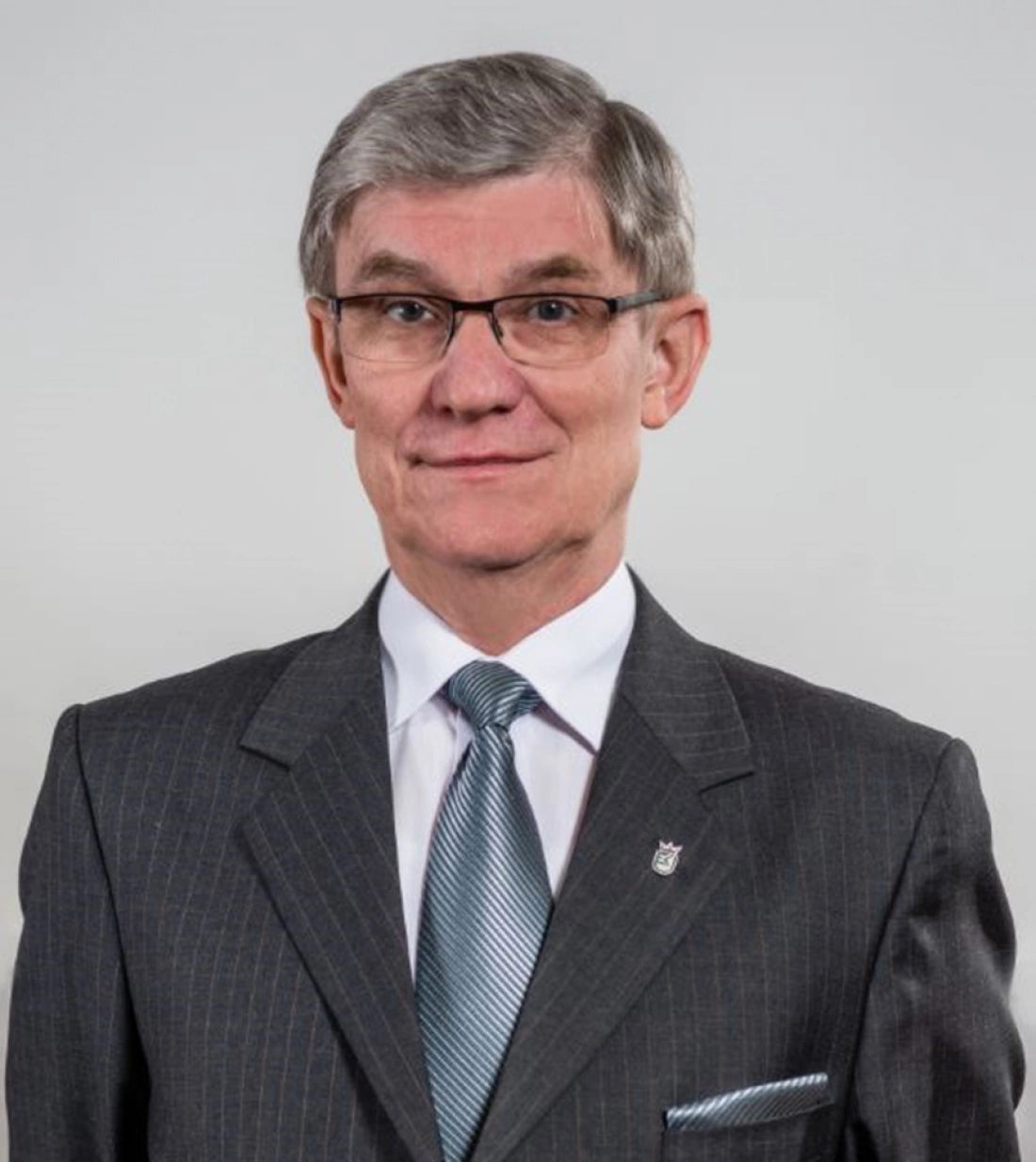 Mężczyzna w eleganckim garniturze z krawatem, noszący okulary, patrzy w stronę kamery.