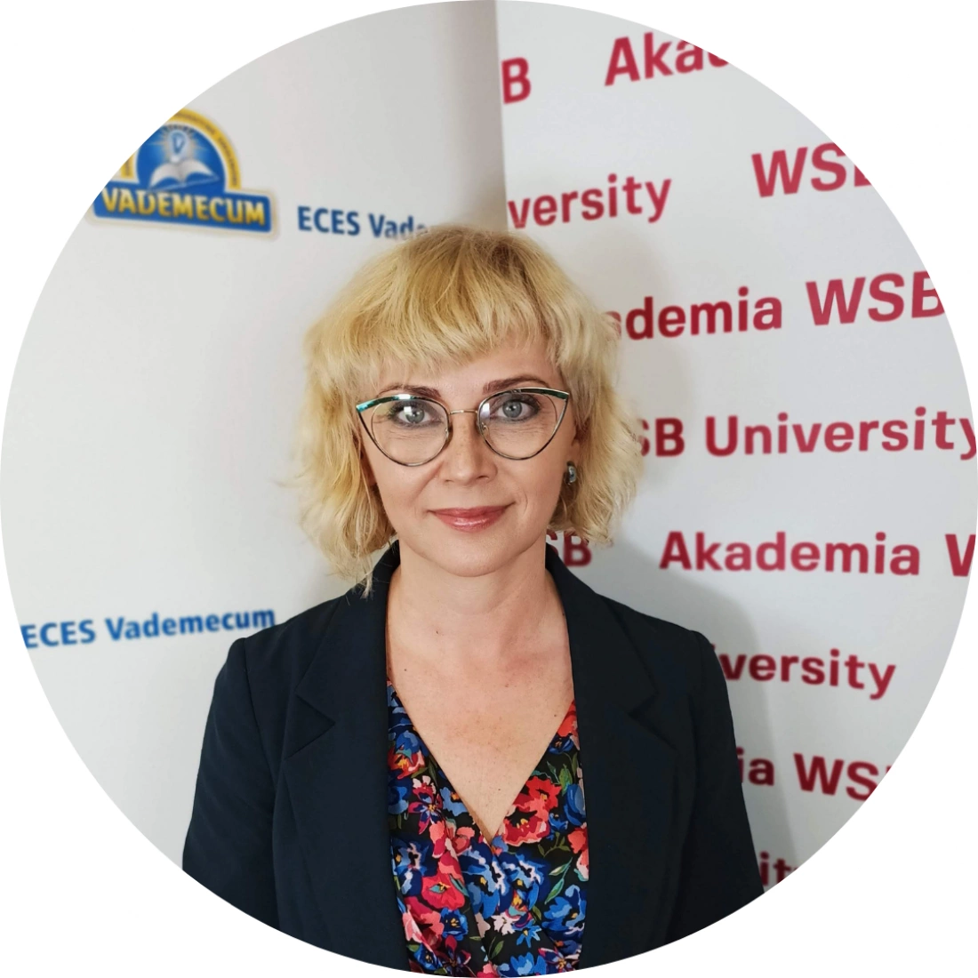 Kobieta z blond kręconymi włosami, w okularach, ubrana w kolorową sukienkę i ciemną marynarkę, stoi przed tłem z logo uczelni.