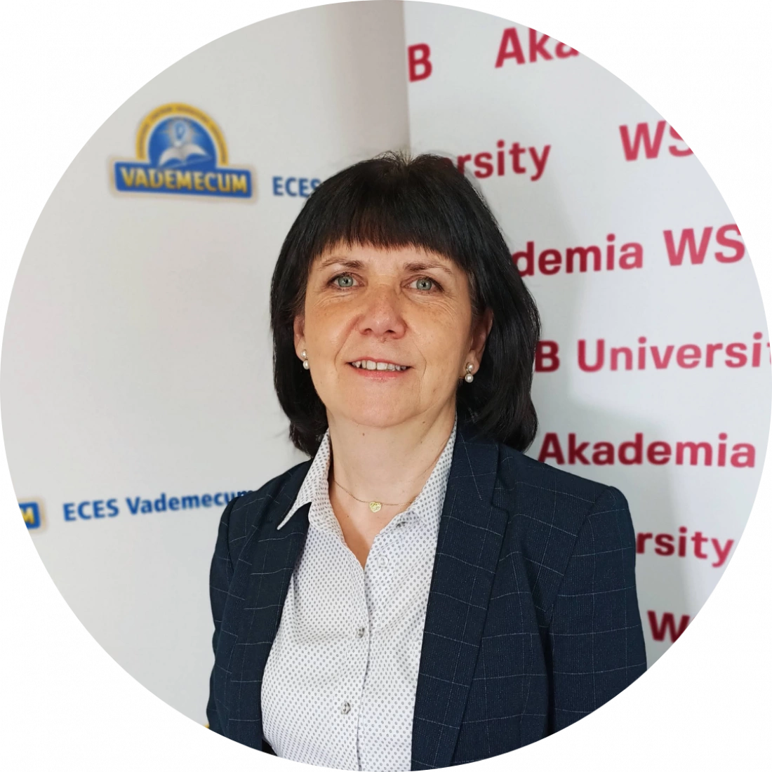 Kobieta w eleganckim stroju, pozująca w biurze z logo uczelni w tle.