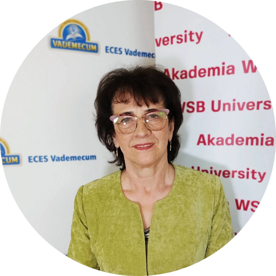 Kobieta w zielonym żakiecie z okularami, stojąca przed tłem z logo uniwersytetu.