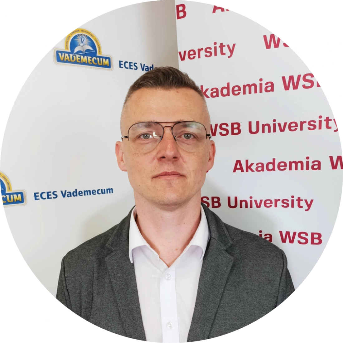 Mężczyzna w okularach, ubrany w szary żakiet, stoi przed tłem z logo Akademii WSB.