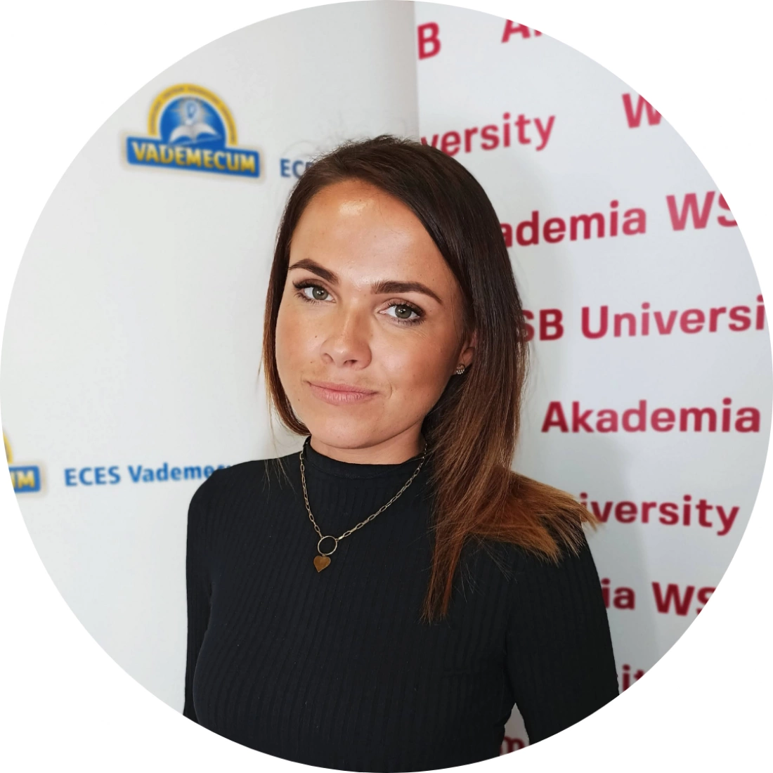 Kobieta z długimi włosami, w czarnej bluzce, stoi przed tłem z logo uczelni.