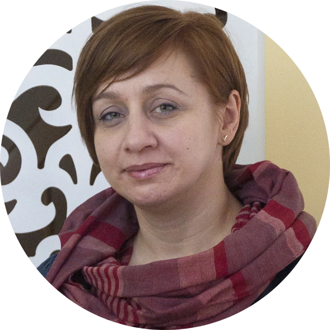Kobieta w czerwono-różowym szaliku, z neutralnym wyrazem twarzy, na tle dekoracyjnej ściany.