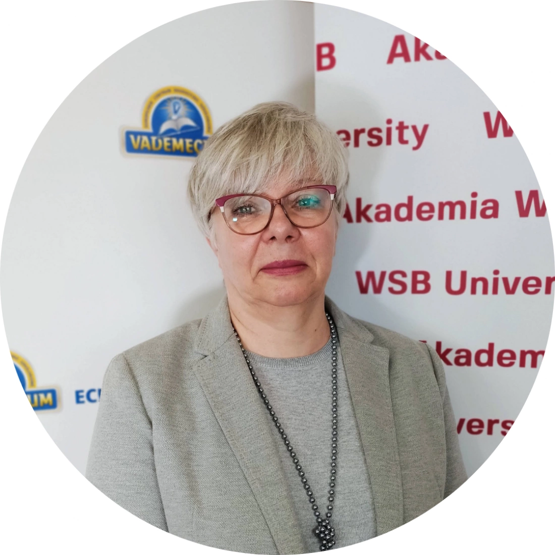 Kobieta z krótkimi, siwymi włosami stoi na tle logotypów akademickich.