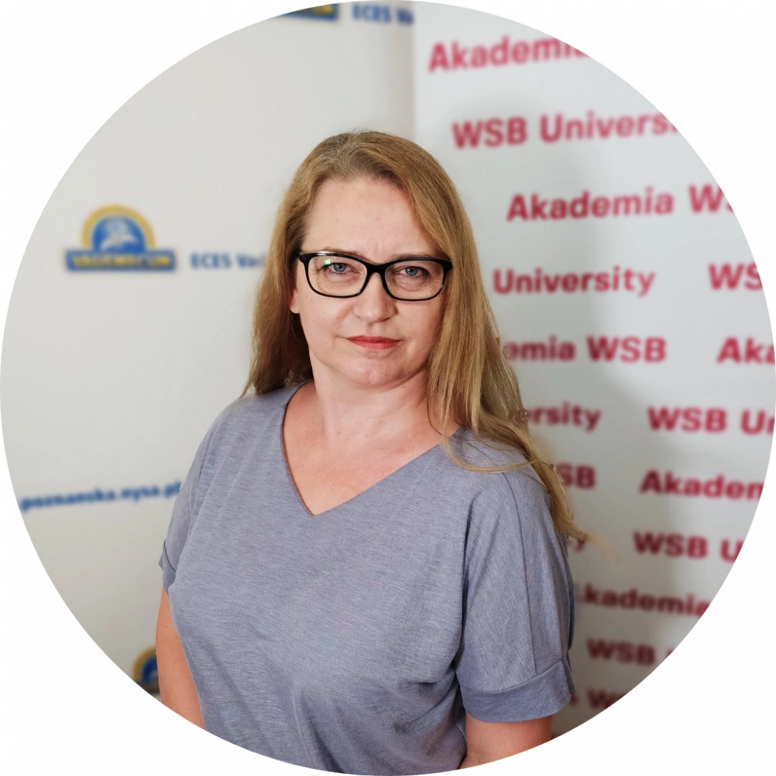 Kobieta w okularach, ubrana w szary t-shirt, stoi przed logo uczelni.