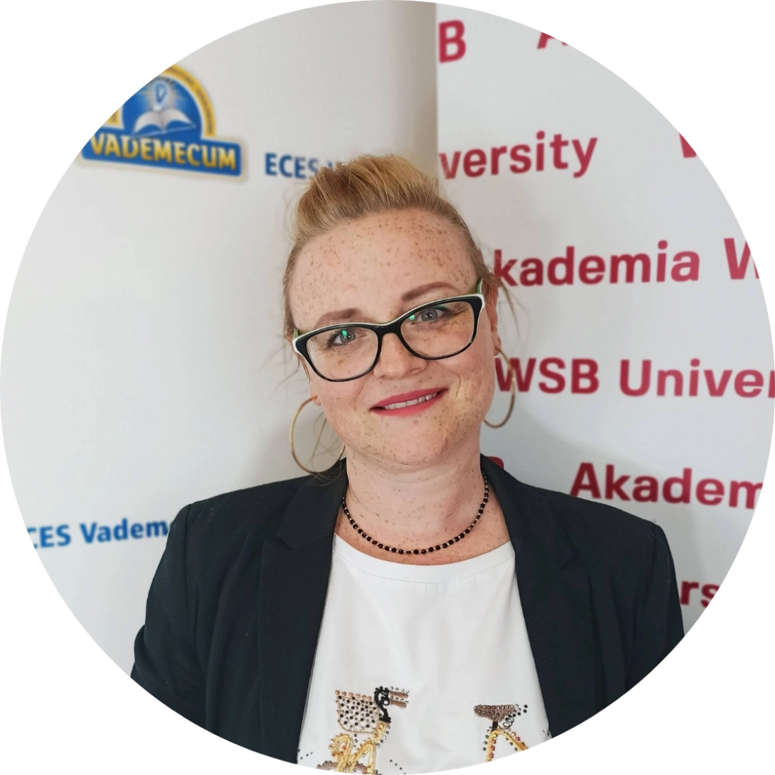Kobieta z rudymi włosami i okularami, uśmiechnięta, stojąca przed tłem z logo uniwersytetu.