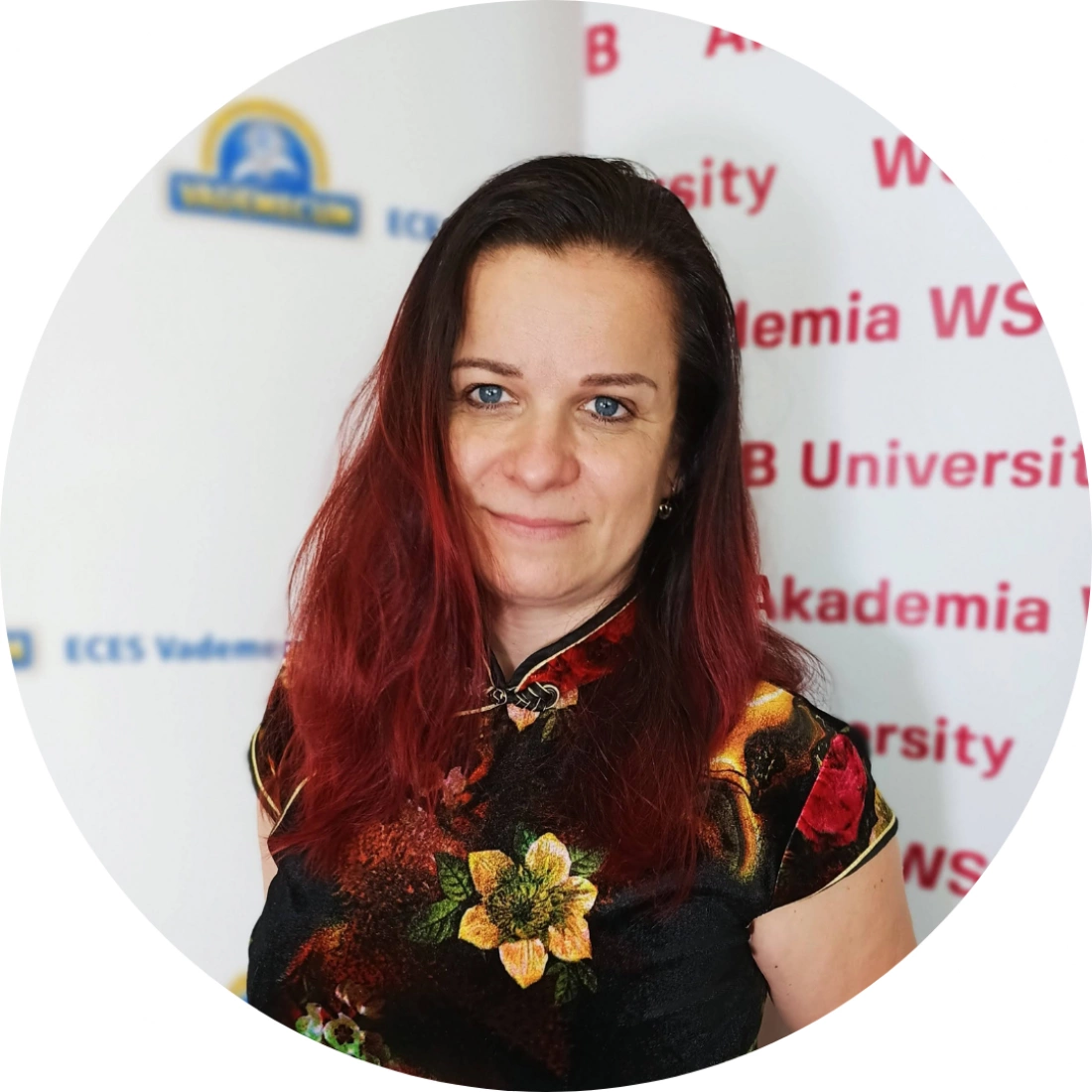 Kobieta w kolorowej sukience z kwiatowym wzorem, pozująca na tle z nazwami uczelni.