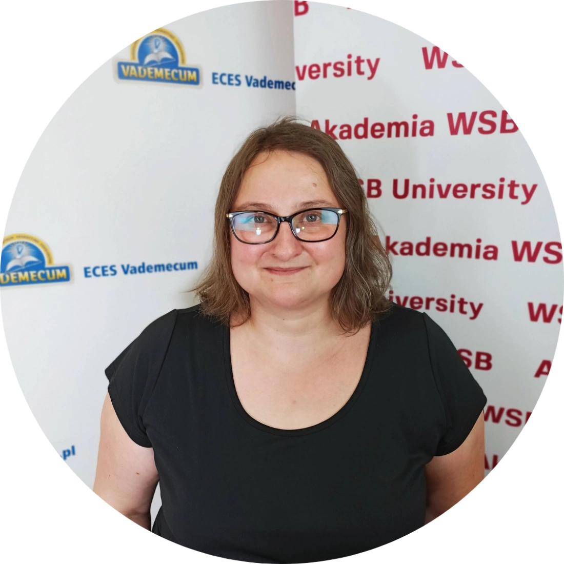 Kobieta w okularach stoi przed banerami Akademii WSB i ECES Vademecum, uśmiechając się do obiektywu.