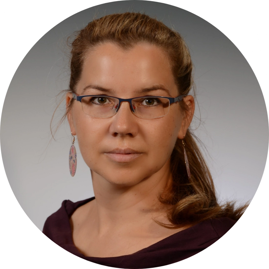Portret kobiety w okularach, z długimi włosami i kolczykami, w jasnym tle.