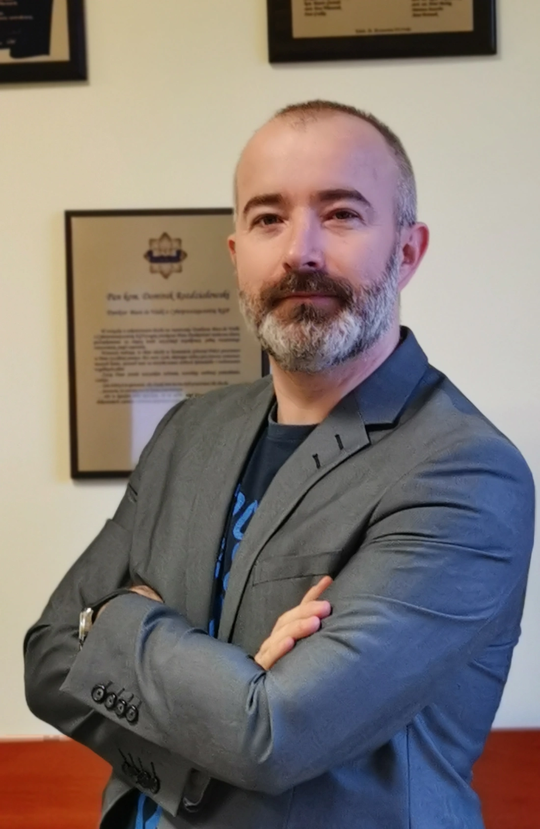 Mężczyzna w szarym garniturze z zarostem, stojący z założonymi rękami na tle ściany z dyplomami.