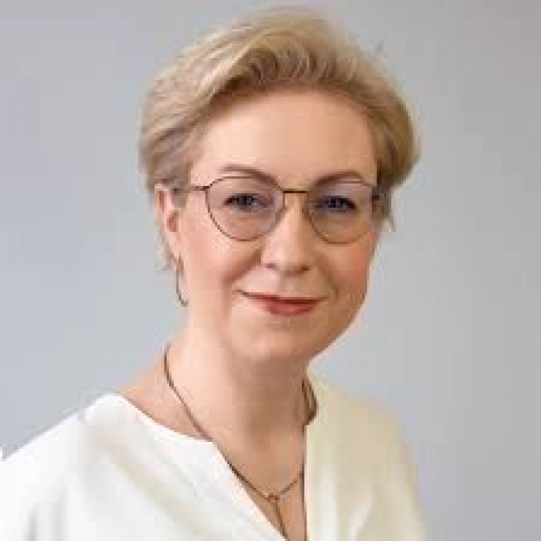 Kobieta z blond włosami, okularami i jasną bluzką, uśmiechająca się do aparatu.
