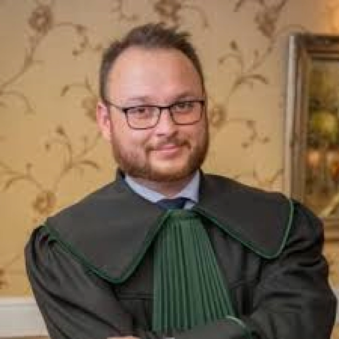 Mężczyzna w togę prawniczą, z okularami, uśmiecha się do kamery.