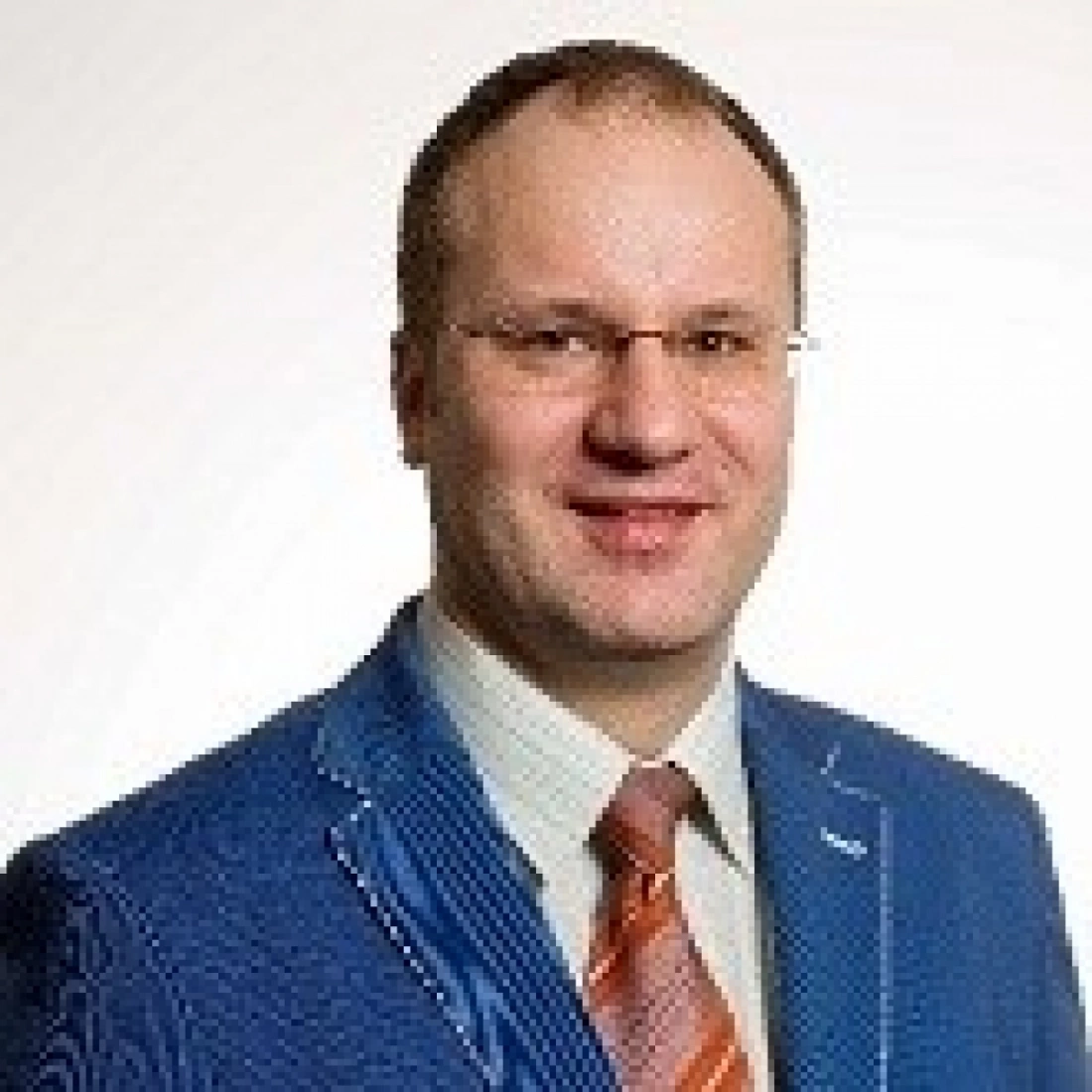 Mężczyzna w niebieskim garniturze z brązowym krawatem, uśmiechający się na tle jasnym.