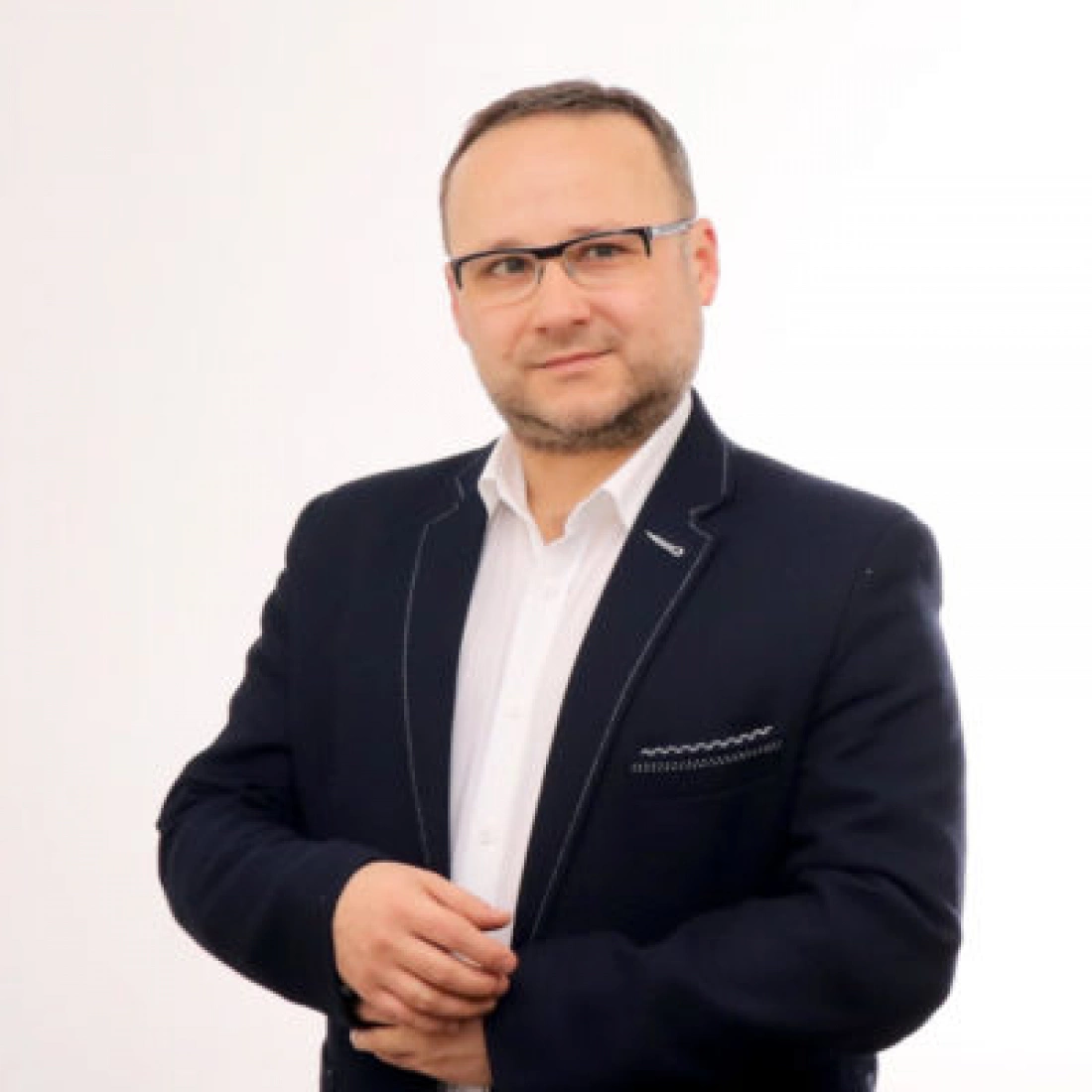 Mężczyzna w eleganckim garniturze, stojący z założonymi rękami na tle białej ściany.