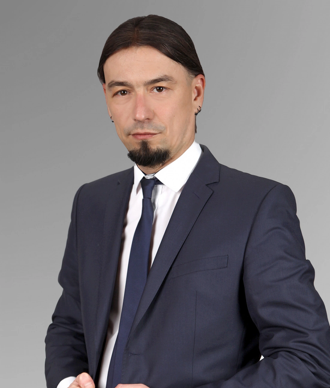 Mężczyzna w garniturze, z długimi, prostymi włosami i piercingiem, stoi w pozie z rękami skrzyżowanymi.