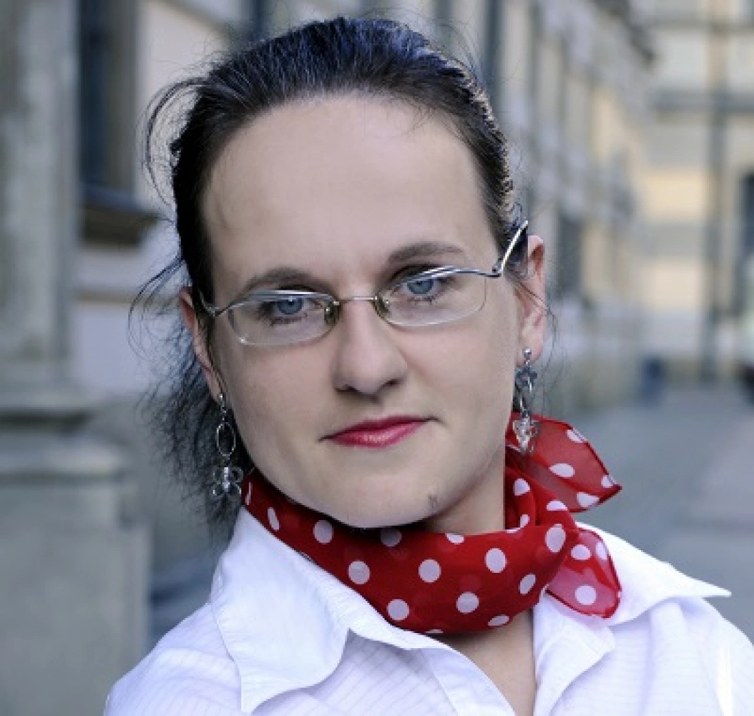 Kobieta w białej koszuli z czerwoną chustą w białe kropki, z okularami i uśmiechem.