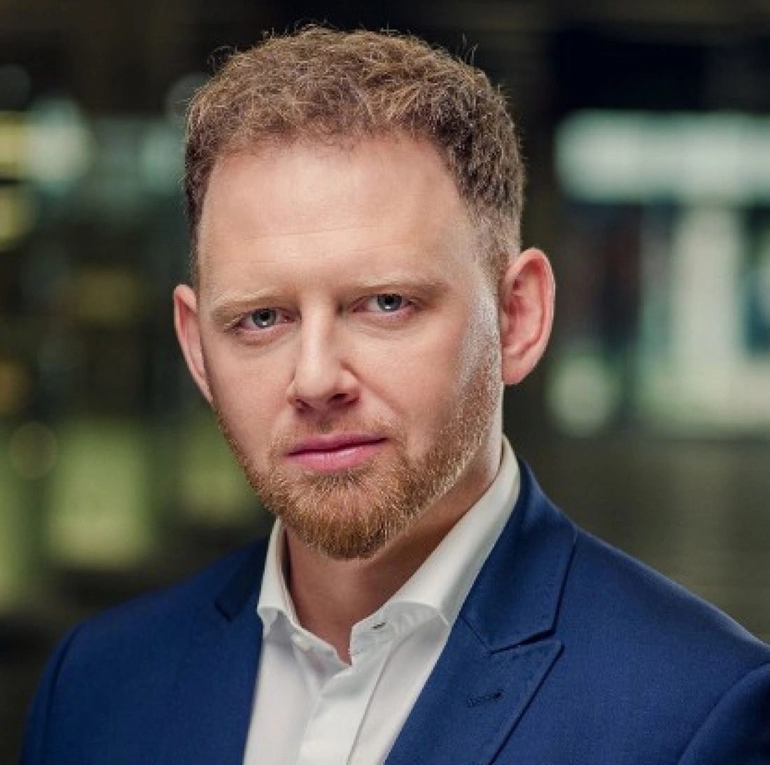 Mężczyzna w eleganckim garniturze z krótkimi, kręconymi włosami i zarostem.