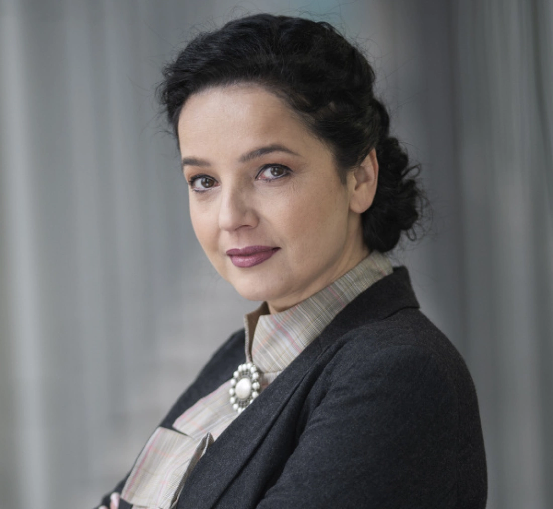 Kobieta w eleganckim stroju, z ciemnymi włosami i delikatnym makijażem, stoi z lekkim uśmiechem na tle jasnej tkaniny.