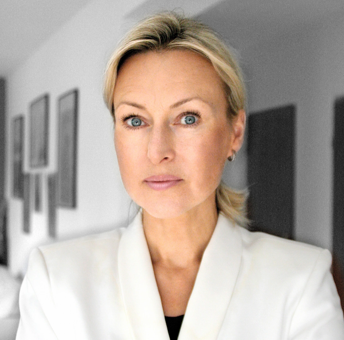 Kobieta w białej marynarce z jasnymi włosami, ujęta z bliska, z poważnym wyrazem twarzy. W tle widać nieostre obrazy na ścianie.