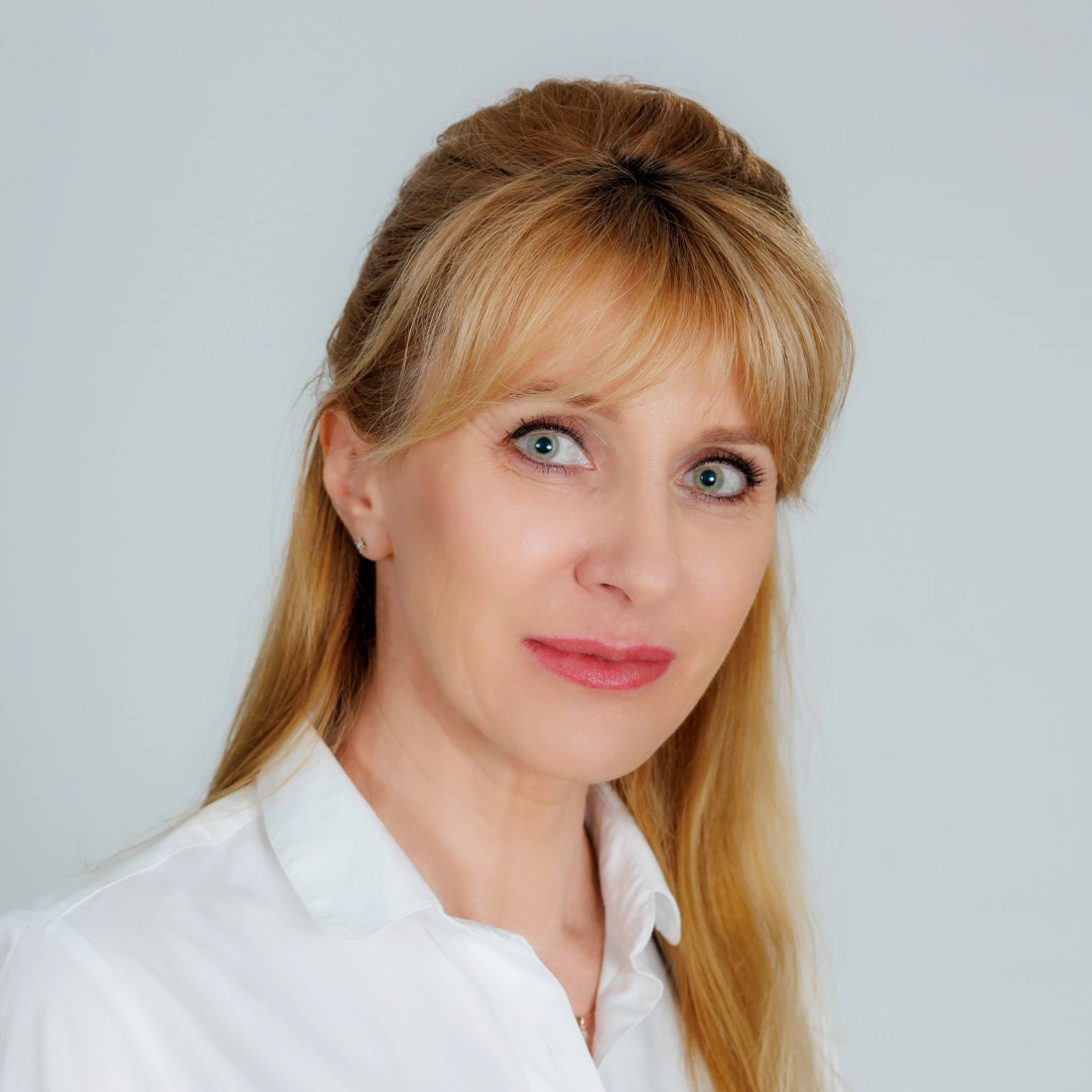 Portret uśmiechniętej kobiety z długimi blond włosami w białej koszuli na neutralnym tle.