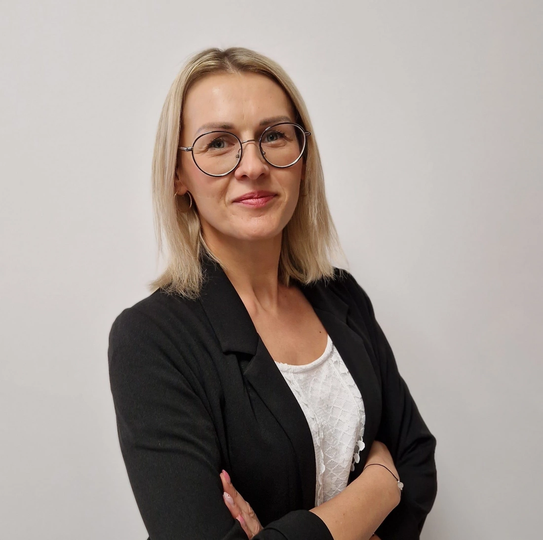 Kobieta z blond włosami, w okularach i czarnym żakiecie, stoi z założonymi rękami na tle jasnej ściany.