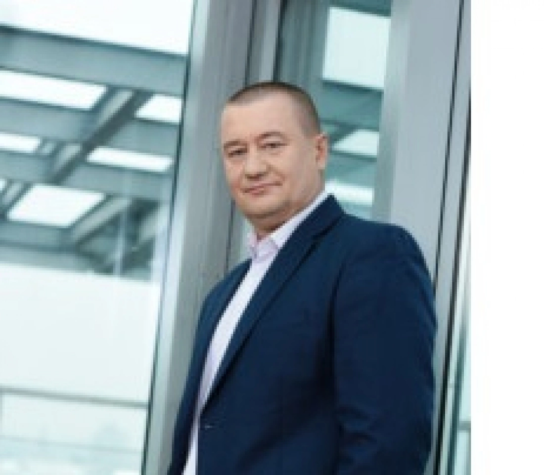 Mężczyzna w garniturze stoi obok szklanej ściany, uśmiechając się do kamery.