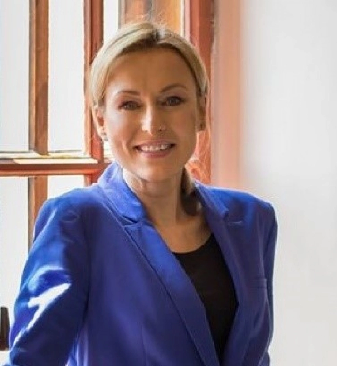 Kobieta w niebieskim żakiecie uśmiecha się przy oknie.